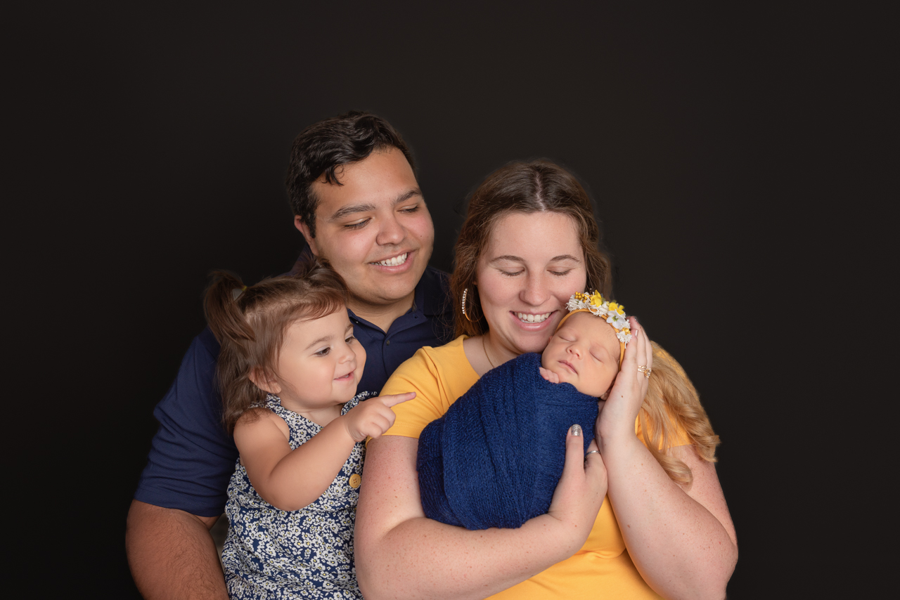 Family of four with toddler smiling at newborn baby wrapped in navy at Ogden Utah photography studio