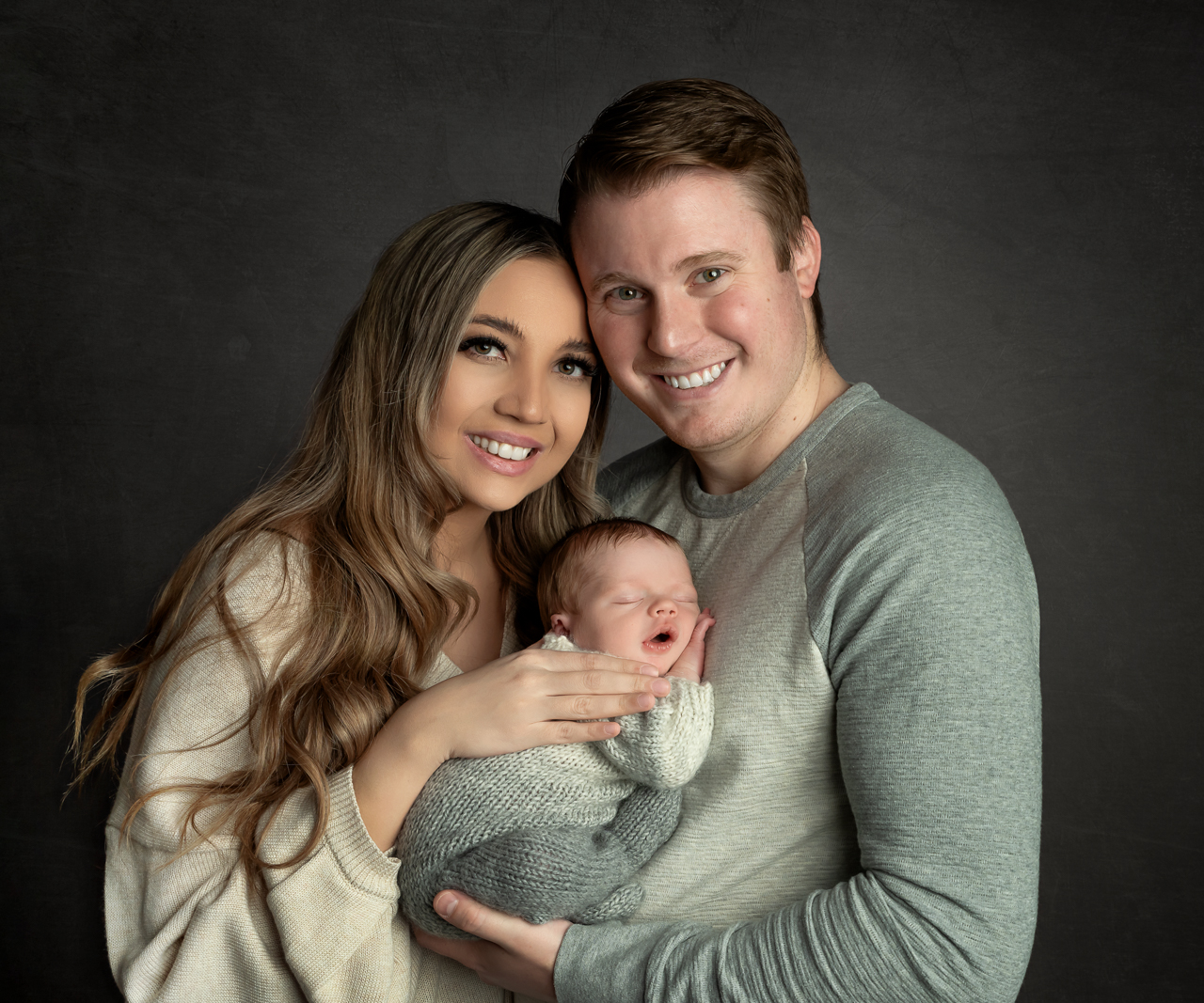 New parents smiling while holding yawning baby in gray wrap at La Bella Sole Photography studio