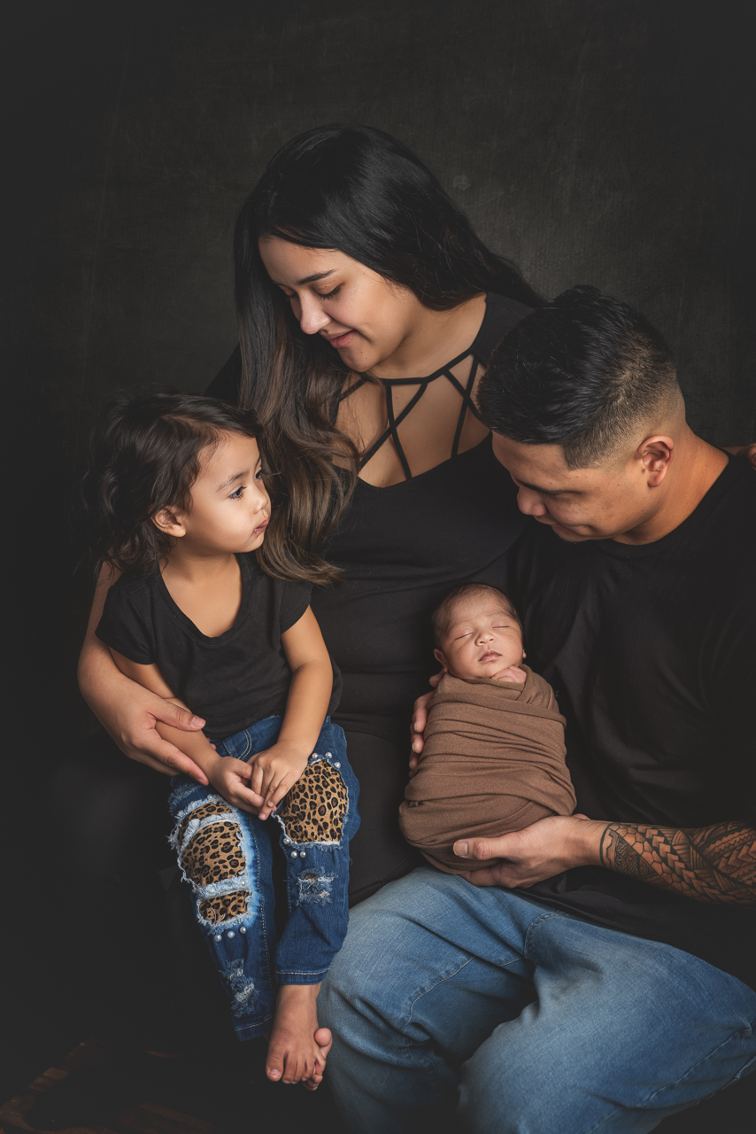 Family of four with toddler sitting next to newborn swaddled in brown during studio family newborn session