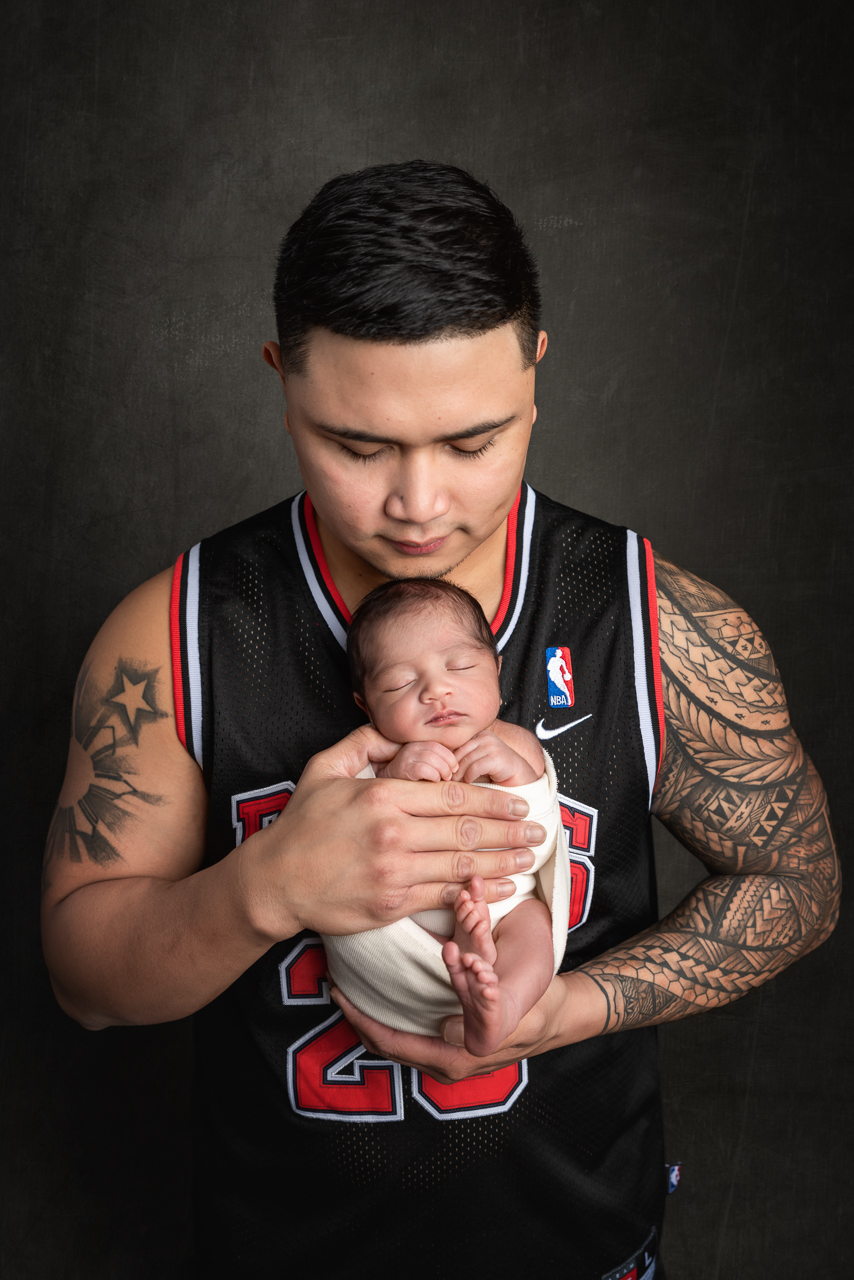 Tattooed father in basketball jersey cradling newborn wrapped in white at North Ogden photo studio