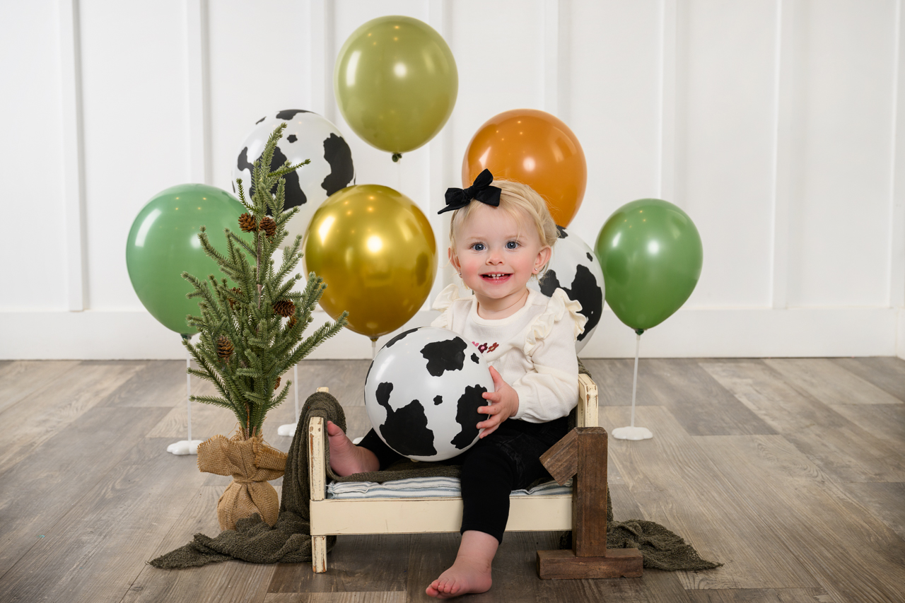 DS1_6288 smash cake holy cow First birthday portrait of smiling baby girl holding cow print balloon in a rustic farm-themed smash cake session at a North Ogden Utah photo studio.