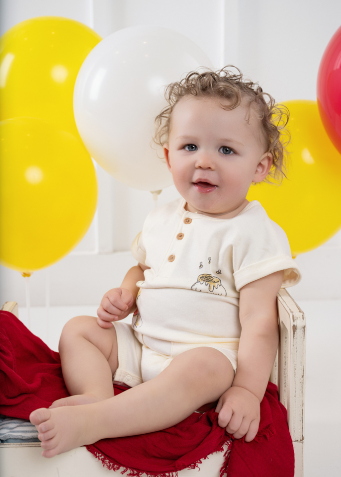 Studio birthday portrait with baby sitting on white bench surrounded by balloons