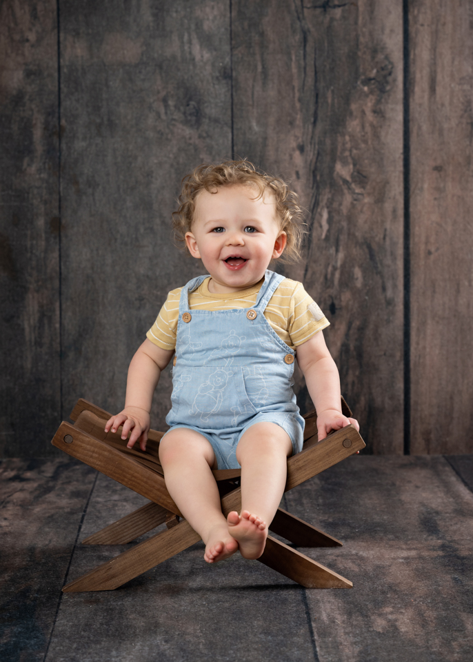 Classic first birthday portrait with baby smiling on rustic wooden chair in front of wood backdrop