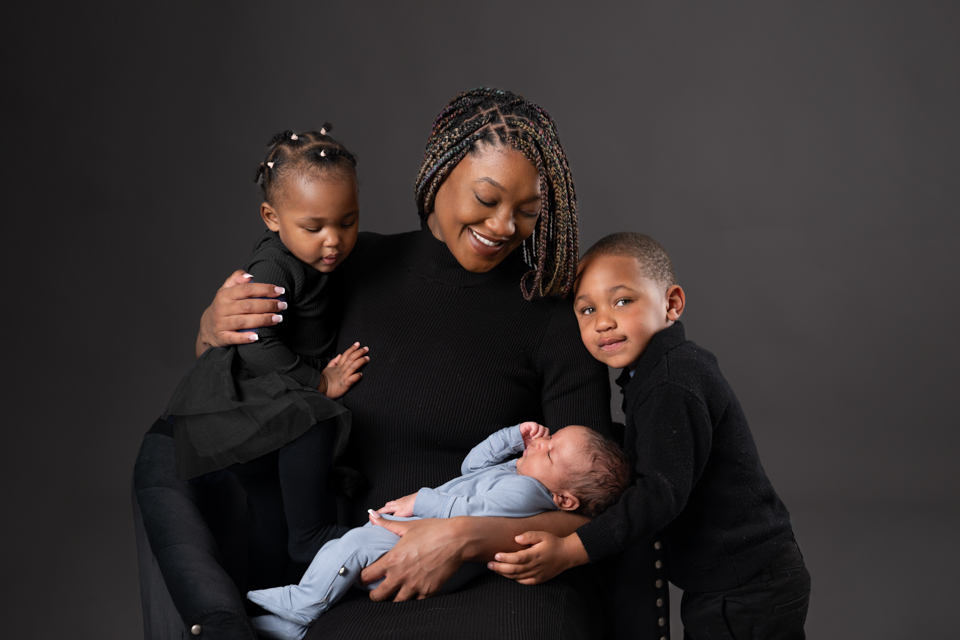 Mother with three children during a newborn studio photography session in North Ogden, Utah, wearing coordinated black outfits on a dark backdrop