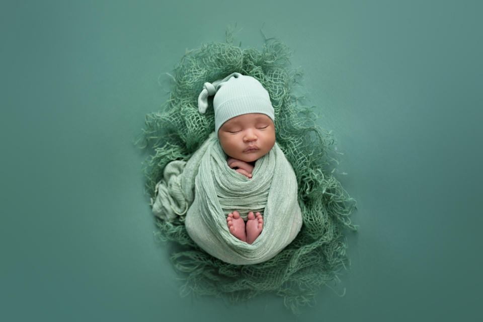 Newborn baby wrapped in soft green fabric and matching hat, peacefully sleeping on a textured green blanket during a studio newborn photography session in North Ogden, Utah